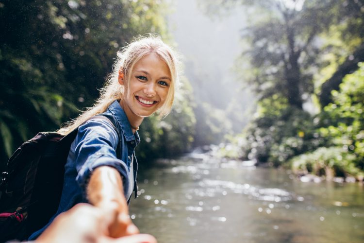 Portrait of happy young woman holding hand of her boyfriend while walking by mountain stream. Couple enjoying a hike in nature.